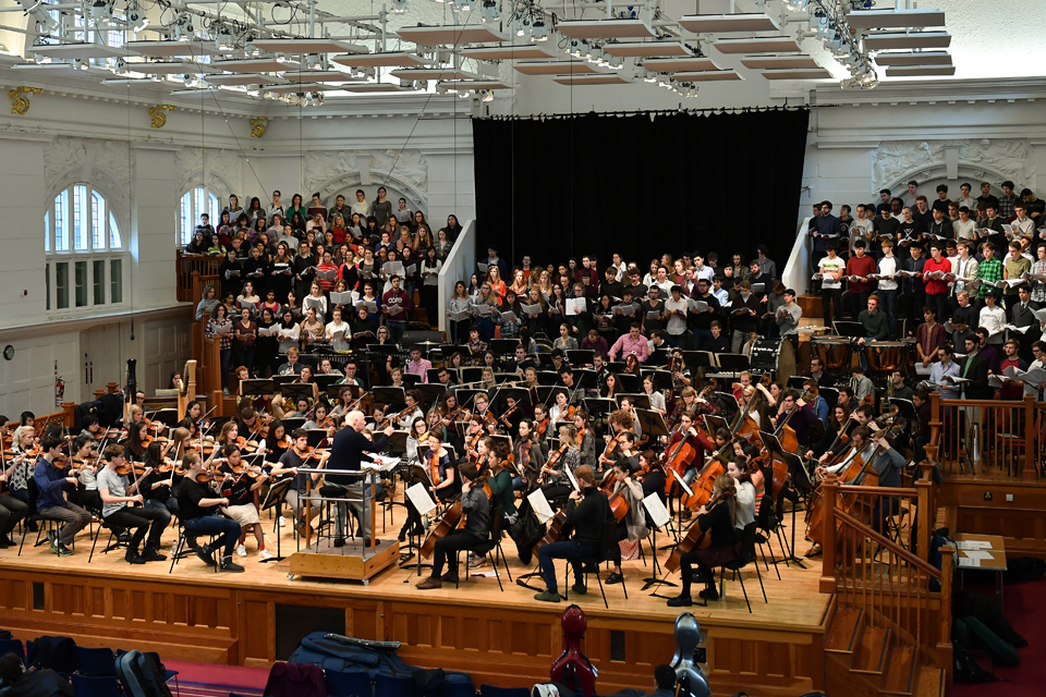 A wide shot of an orchestra rehearsing, located on a stage, being conducted by a older man with white hair, with choir groups in the back of the stage.
